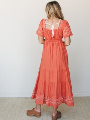 A warm terracotta midi dress shot from behind, showing off its embroidered hem and sleeves, open tie-back detail, and tiered skirt — paired with wood-platform clogs for that effortlessly earthy, boho-everyday feel.