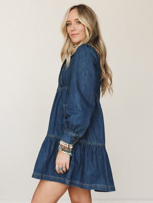 A denim mini dress with boho soul — the tiered ruffle hem and gathered long sleeves give it that lived-in, effortless feel, styled with stacked beaded bracelets and turquoise rings for a personal touch.