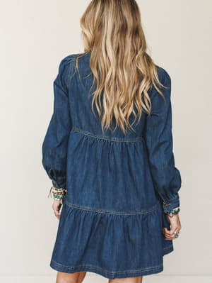 A tiered dark denim mini dress shot from the back, with billowy long sleeves and relaxed gathering that gives it a free-spirited, effortless feel — styled with layers of turquoise and mixed-metal bracelets that say 'I put thought into this, but make