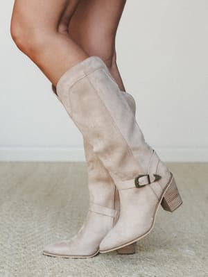 A close-up lifestyle shot of a knee-high suede western boot in warm sand, styled with a buckle strap detail and stacked heel — the kind of boot that makes any outfit feel like it has a story.