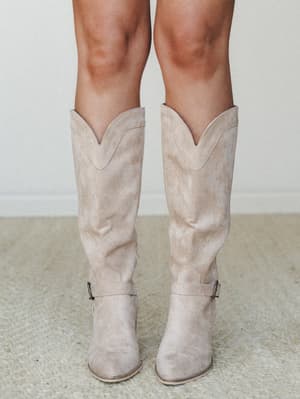 A close-up of knee-high western-style boots in a soft beige suede, styled on a model against a neutral interior backdrop — the kind of boots that do the heavy lifting for any outfit.