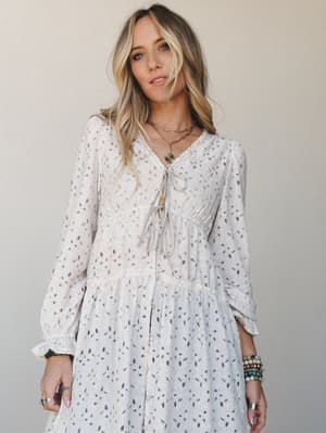 A flowy long-sleeve dress in soft oatmeal with scattered metallic dash print, styled with layered gold necklaces and stacked boho bracelets — the kind of dress that feels like a hug and looks like intention.