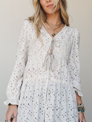 A dreamy long-sleeve dress in soft oatmeal, shot close against a neutral studio wall — the kind of piece that looks like it was made for golden-hour wandering, styled with layered gold necklaces and stacked turquoise bracelets.