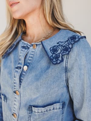 A close-up of a light wash denim jacket with a oversized peter pan collar adorned with dark navy floral rose appliqué, snapped shut with gold hardware — the kind of piece that makes a plain outfit feel like a whole personality.