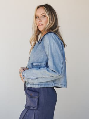 A side-profile studio shot of a model in a washed light blue cropped denim jacket layered over navy cargo pants, styled with stacked rings — easy, relaxed, and put-together all at once.