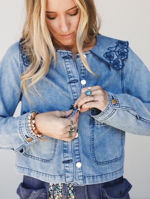 A blonde model looks down while buttoning a light-wash denim jacket with floral embroidery on the collar, her hands stacked with silver and turquoise rings and beaded bracelets — the kind of jacket that does all the talking.
