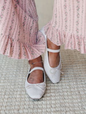 A close-up of white quilted ballet flats with ankle straps and bow detail, paired with a blush floral ruffle-hem maxi dress — soft, feminine, and the kind of outfit that makes a Saturday feel intentional.