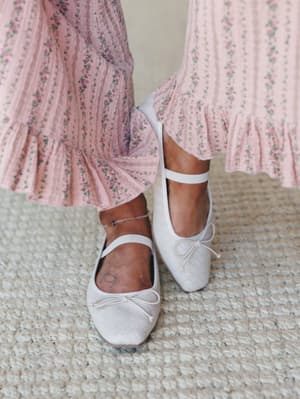 A close-up of white quilted ballet flats with ankle straps and bow detail, paired with a blush floral ruffle-hem maxi dress — soft, feminine, and the kind of outfit that makes a Saturday feel intentional.