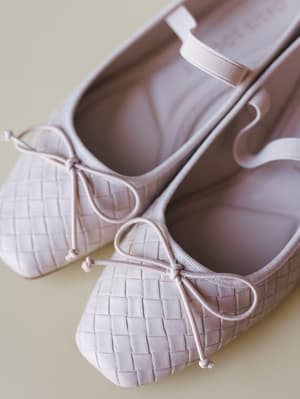 A close-up of blush-beige woven ballet flats with delicate bow ties at the toe, shot against a soft sage-green surface — the kind of shoe that makes every outfit feel like it was planned.