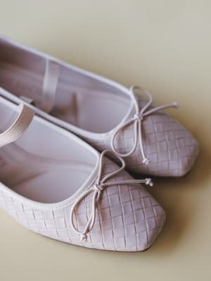 A pair of blush-toned woven ballet flats with delicate bow ties rest against a soft yellow-green surface — the kind of shoes you buy and immediately want to wear everywhere.