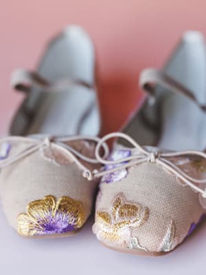 A close-up of two pairs of floral-embroidered ballet flats — one in warm sand linen, one in soft sage — arranged toe-to-toe against a dusty pink backdrop, with the intricate gold and lavender embroidery doing all the talking.