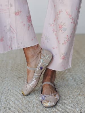A close-up of embroidered floral ballet flats with ankle strap detailing, styled against wide-leg floral print pants and a delicate gold anklet — soft, romantic, and quietly considered.