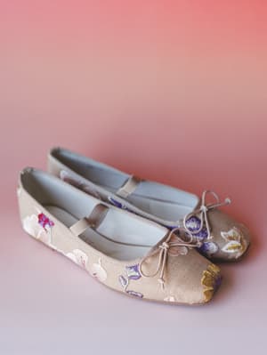 A pair of embroidered ballet flats in warm taupe sit against a soft pink gradient background — the kind of shoe that looks equally at home on a brunch patio or a garden wedding.