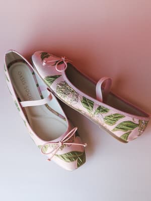 A pair of blush brocade ballet flats with green leaf and rose jacquard detailing, finished with dainty bow ties at the toe — shot overhead on a soft pink gradient background that feels like the inside of a flower.