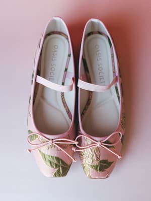 A pair of brocade Mary Jane flats shot from overhead against a soft pink backdrop — the kind of shoes that make you rethink every outfit you already own.