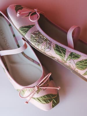 A close-up of a pair of pink brocade ballet flats with floral jacquard detailing, bow tie closures, and elastic ankle straps — the kind of shoe that makes you feel like you planned your whole outfit around them.