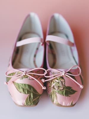 A pair of pink brocade Mary Jane flats with floral leaf detailing and delicate bow ties, shot close-up against a soft rose-toned background — the kind of shoes that feel like a secret.