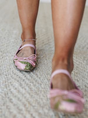 A close-up shot of pink floral ballet flats with ankle straps mid-stride on a woven jute rug — the kind of shoe that makes a whole outfit feel intentional without trying too hard.