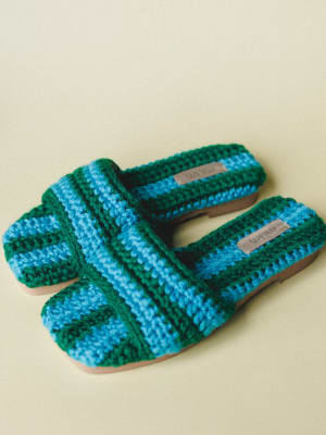 A pair of chunky crochet slide sandals in bold blue and forest green stripes, shot close-up against a soft cream background — the kind of shoes that make a whole outfit feel more intentional.