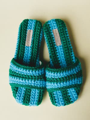 A pair of chunky crochet slide sandals in bold blue and green stripes, shot flat against a warm cream background — the kind of shoes that make you want to pack a beach bag.