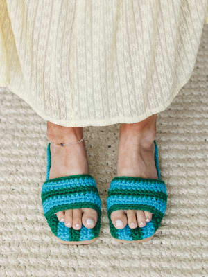 A close-up shot from above of crochet slide sandals in bold blue and green stripes, worn with a cream lace midi skirt and a delicate anklet — the kind of detail shot that makes you want to zoom in and stay there.