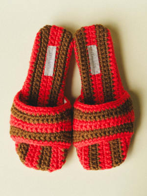 A pair of chunky crochet slide sandals in bold red and walnut brown stripes, shot overhead against a soft cream background — the kind of shoes that make you want to repaint your toenails and book a trip somewhere.