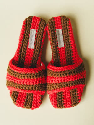 A pair of chunky crochet slide sandals in bold red and walnut brown stripes, shot overhead against a soft cream background — the kind of shoes that make you want to repaint your toenails and book a trip somewhere.
