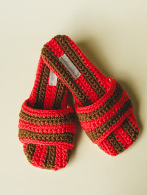 A pair of chunky crochet slide sandals in bold red and brown stripes, shot flat against a soft cream background — the kind of shoes that make you want to pad around the house like it's a lifestyle.