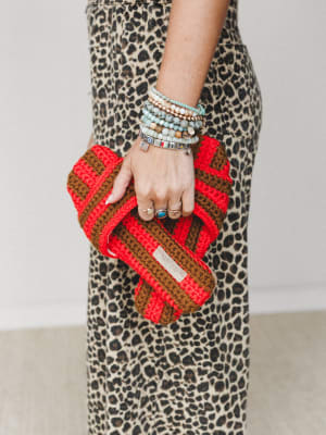 A close-up of a model's hand clutching a chunky crochet clutch in bold red and brown stripes, layered against a leopard print skirt — the bracelet stack and stacked rings doing just as much styling work as the bag itself.