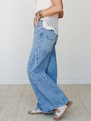 A side-profile shot of wide-leg barrel jeans in a medium wash denim, paired with a cream crochet top, stacked beaded bracelets, and white buckle sandals — the kind of effortless outfit that looks like it took five minutes and somehow works perfectly.