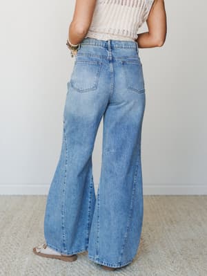 Shot from behind, a model wears roomy wide-leg barrel jeans in a faded medium wash denim, paired with a cream open-knit top and strappy sandals — the kind of effortless, lived-in outfit you reach for on a good day.
