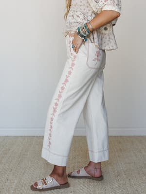 A side-profile shot of wide-leg cream pants with a trailing pink floral embroidery detail running down the leg, styled with a lace and floral top, stacked turquoise bracelets, and neutral buckle sandals — soft, expressive, and very much a Saturday wi