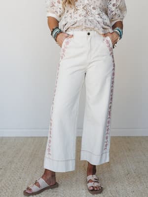 Cream wide-leg pants with delicate pink floral embroidery running along the side seams and hem, styled with a lace top, beaded bracelets, and buckle sandals — the kind of outfit that makes a Saturday feel intentional.