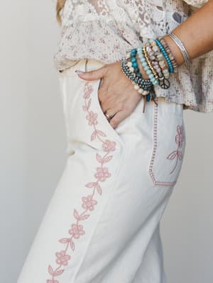 A close-up detail shot of cream wide-leg pants with a trailing pink floral embroidery running down the side seam, styled with a ditsy floral lace-trimmed top and a stack of boho bracelets — the kind of outfit that feels intentional without trying too