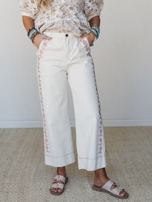 Cream wide-leg pants with delicate pink floral embroidery trailing down the outer seams and along the hem, styled with a lace top, stacked beaded bracelets, and buckle sandals — the kind of outfit that looks effortless but clearly wasn't an accident.