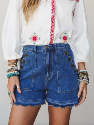 A mid-shot pairing of high-waisted scallop-hem denim shorts with a white embroidered blouse, layered bracelets, and a woven bag strap — the kind of outfit that feels effortless but definitely didn't happen by accident.