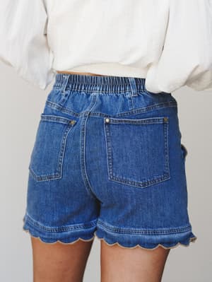 A close-up back view of medium-wash denim shorts with a playful scalloped hem, styled with a white puff-sleeve top — the kind of detail that makes a basic seem anything but.