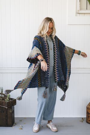 A woman with long blonde hair smiles, wearing the Desert Dreams Crochet Ruana - Cobalt over a light blue jumpsuit and pink sneakers, standing before a white wall with a wooden trunk and potted plant—a perfect boho-chic look.