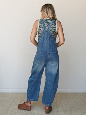 A back-facing model in relaxed, sunwashed denim overalls layered over a camo tank, styled with brown studded clogs — the kind of outfit that feels effortless and a little nostalgic all at once.