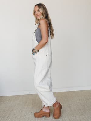 A relaxed side-profile shot of a model in cream wide-leg denim overalls layered over a grey printed tank, finished with wood-heeled clogs and a stack of beaded bracelets — the kind of effortless outfit that somehow looks like it took no effort at all
