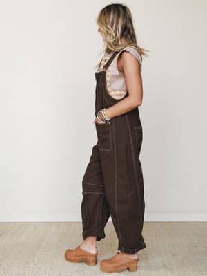 A side-profile shot of a model wearing relaxed dark brown overalls with contrast stitching, layered over a striped tank, finished with stacked beaded bracelets and tan studded clogs — the kind of outfit that looks like it was pulled together effortle