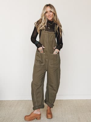 Olive canvas overalls styled over a sheer black long-sleeve top with turquoise jewelry and tan clogs — the kind of effortlessly lived-in look that makes a farmer's market run feel like a whole moment.