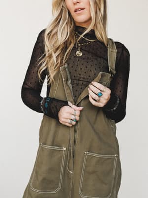 An olive canvas overall dress layered over a sheer black polka dot turtleneck, styled with stacked silver and turquoise rings and layered gold chain necklaces — the kind of put-together-but-effortless look that feels vintage without trying.