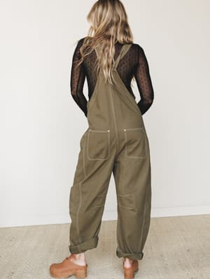 Shot from the back, a model wears roomy olive canvas overalls over a sheer black dotted long-sleeve top, styled with tan wooden clogs — the kind of outfit that feels equally at home on a farmers market Saturday or a casual creative workday.