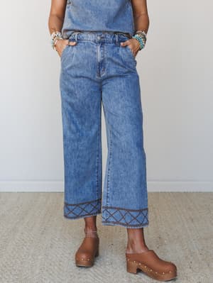 Wide-leg cropped denim pants with contrast brown embroidery at the hem and waistband, styled with wood-platform clogs and a stack of mixed bracelets — earthy, relaxed, and quietly cool.