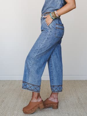 A side-profile shot of wide-leg cropped denim with standout geometric embroidery at the hem, styled with stacked beaded bracelets and warm brown clogs — the kind of jeans that do the talking for you.