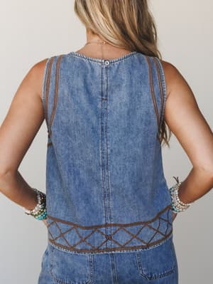 A back-view detail shot of a medium-wash denim top with warm brown embroidered trim and geometric stitching at the hem, styled with stacks of beaded bracelets — equal parts vintage Western and everyday boho.