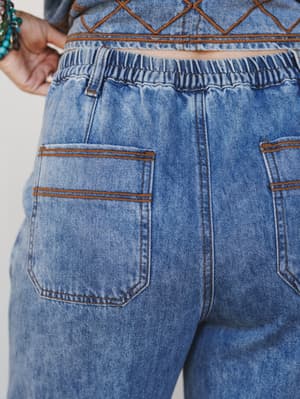 A close-up detail shot of the back of acid-washed denim pants featuring an elastic waistband and patch pockets with distinctive rust-brown contrast stitching, paired with a matching embroidered denim top visible at the crop.