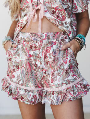 A close-up shot of a paisley-printed two-piece set — ruffle-hem shorts with lace trim and a matching crop top with tie closure — styled with stacked boho bracelets for a free-spirited summer feel.