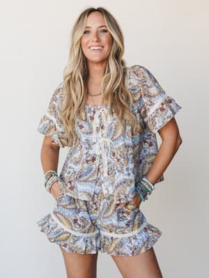 A smiling model in a matching paisley print top and ruffle-hem shorts set in powder blue, styled with stacked turquoise bracelets and a delicate necklace — effortlessly boho and made for a warm-weather day that goes anywhere.